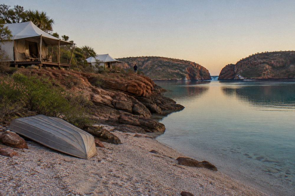 White safari tents on a red dirt coastline above calm turquoise water