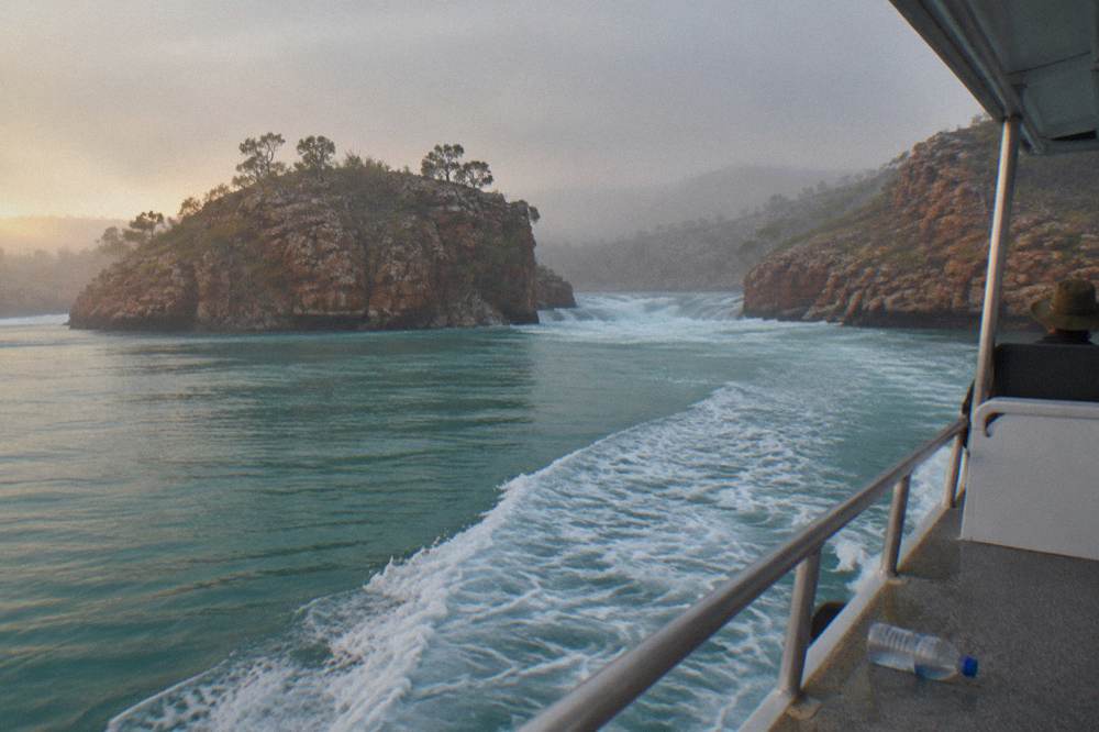 A boat wake crossing a bay where calm water meets a line of churning rapids between rocks
